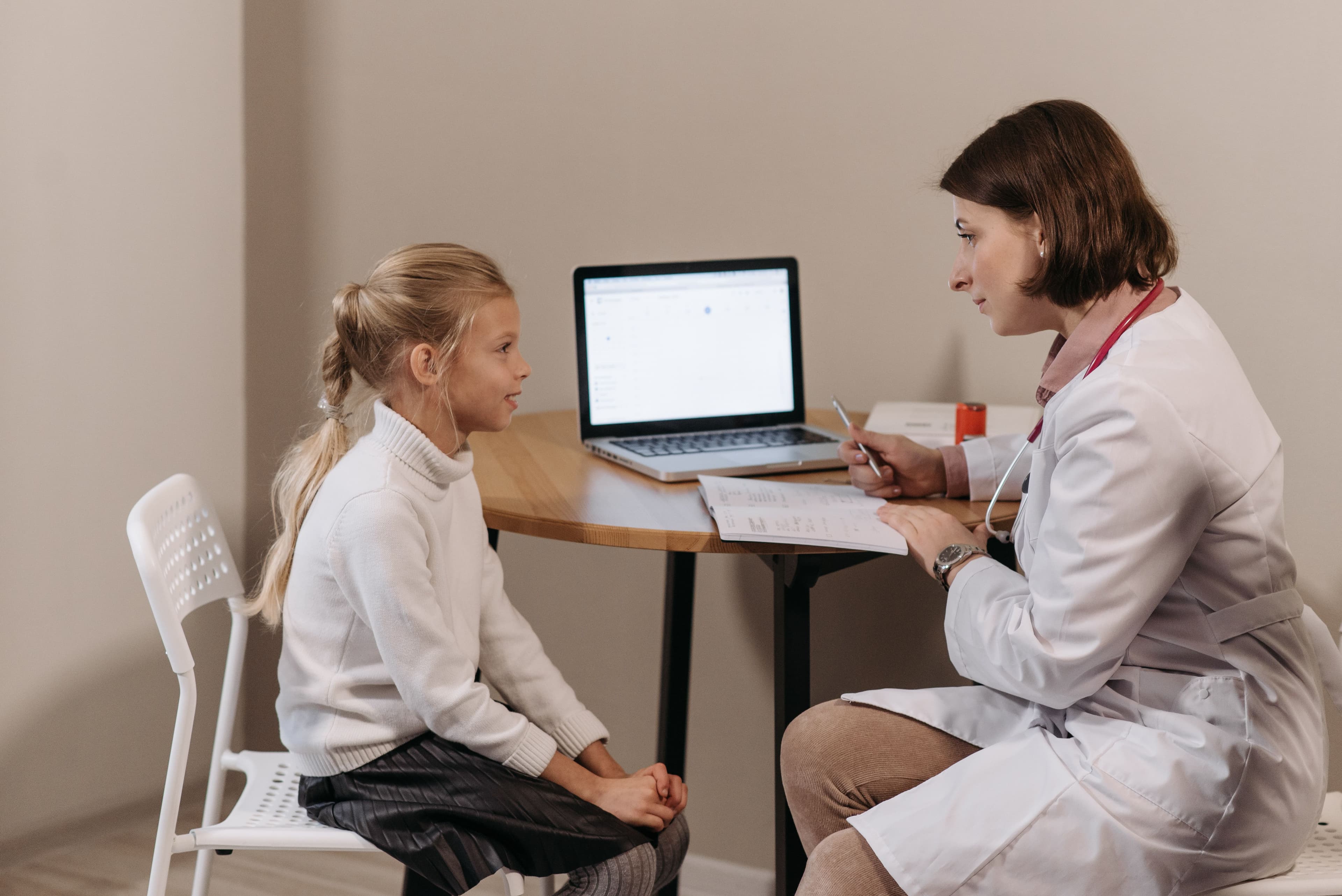 Doctor in lab coat consulting a young girl in a clinic with a laptop on the table