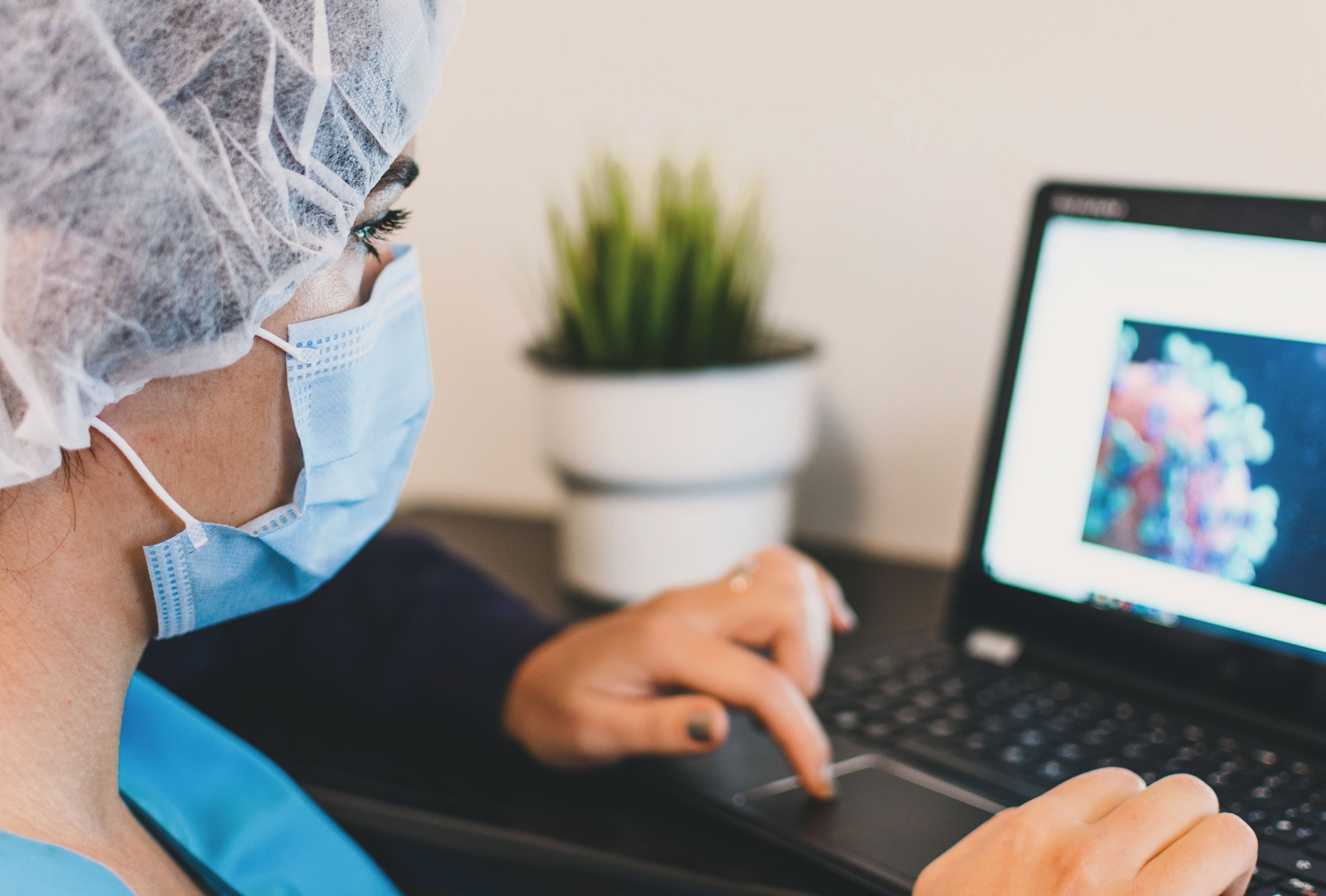 A healthcare worker typing on a laptop while wearing a face mask in an indoor setting