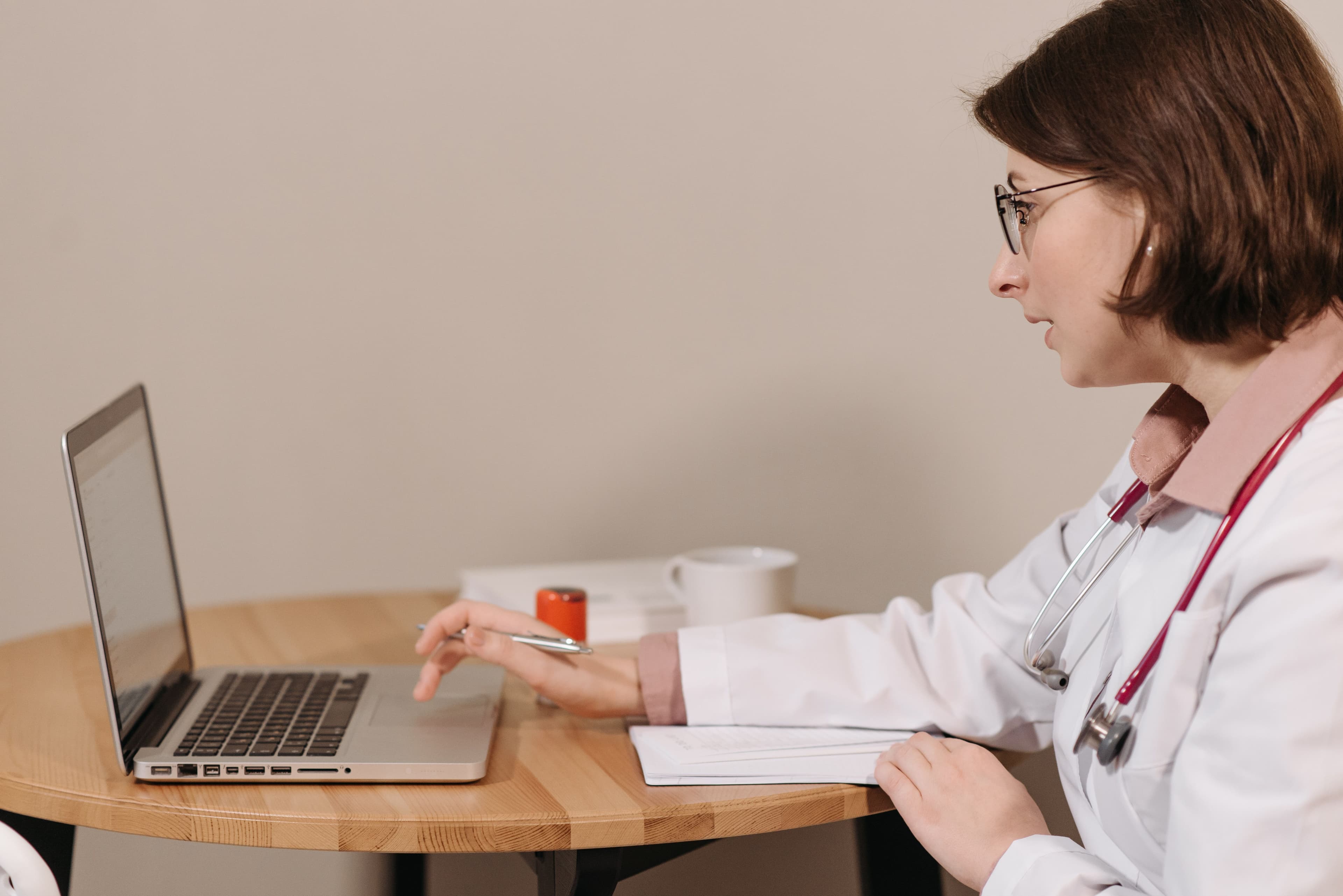 A female doctor in a white coat using a laptop while writing notes at a wooden table