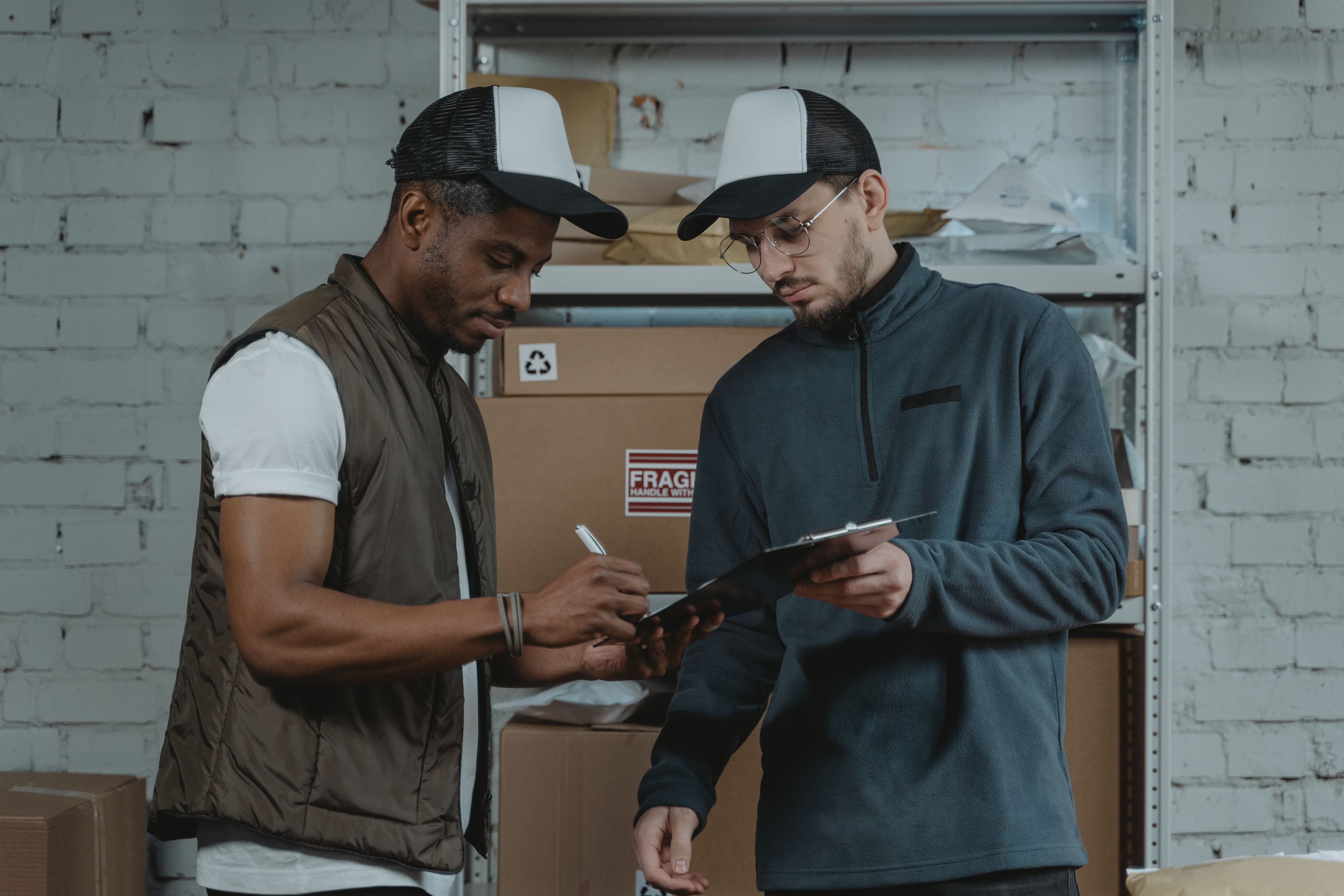 Two male couriers discussing and signing a clipboard in a warehouse setting, surrounded by packages