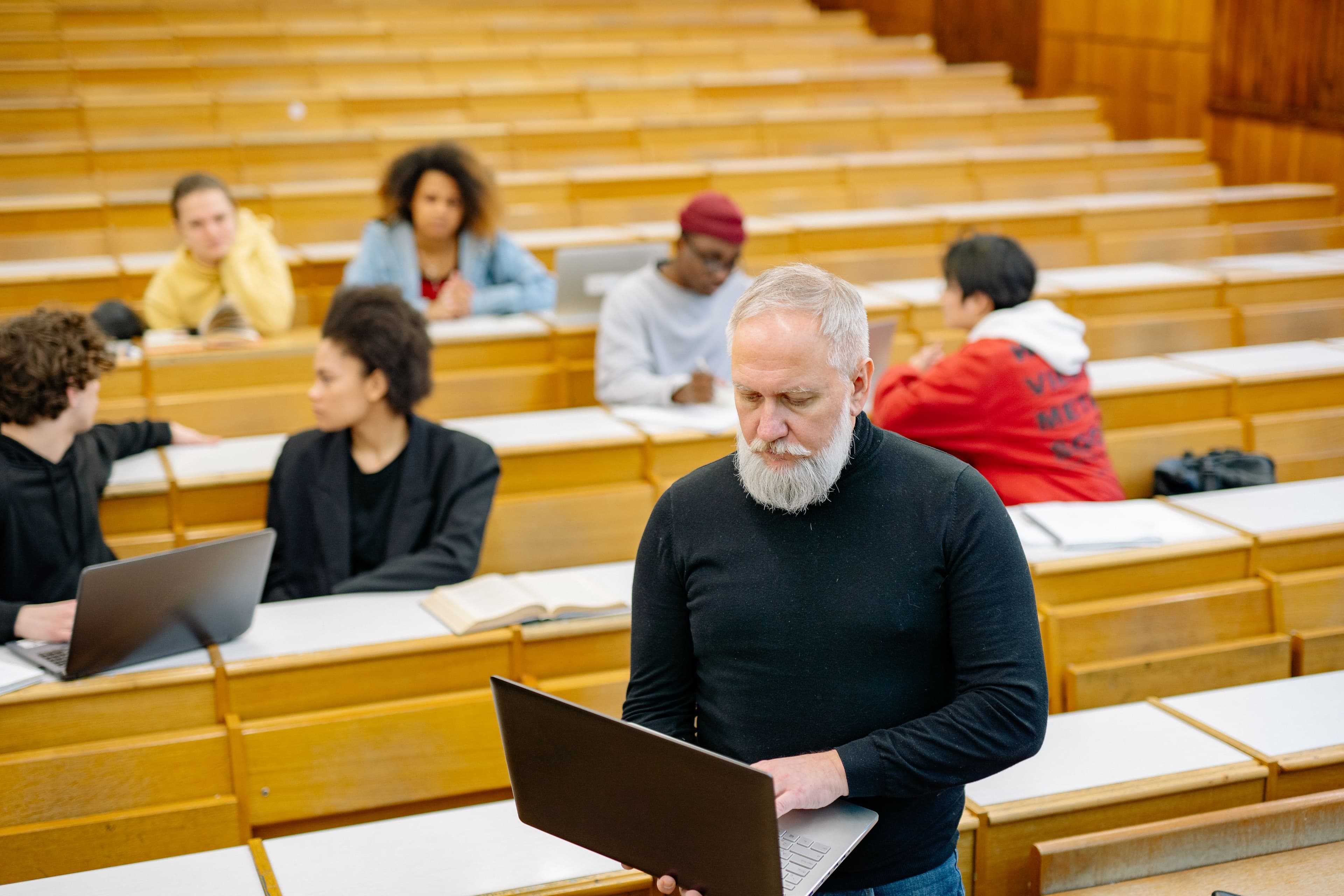 Mature teacher interacting with diverse students using laptops in a spacious lecture hall